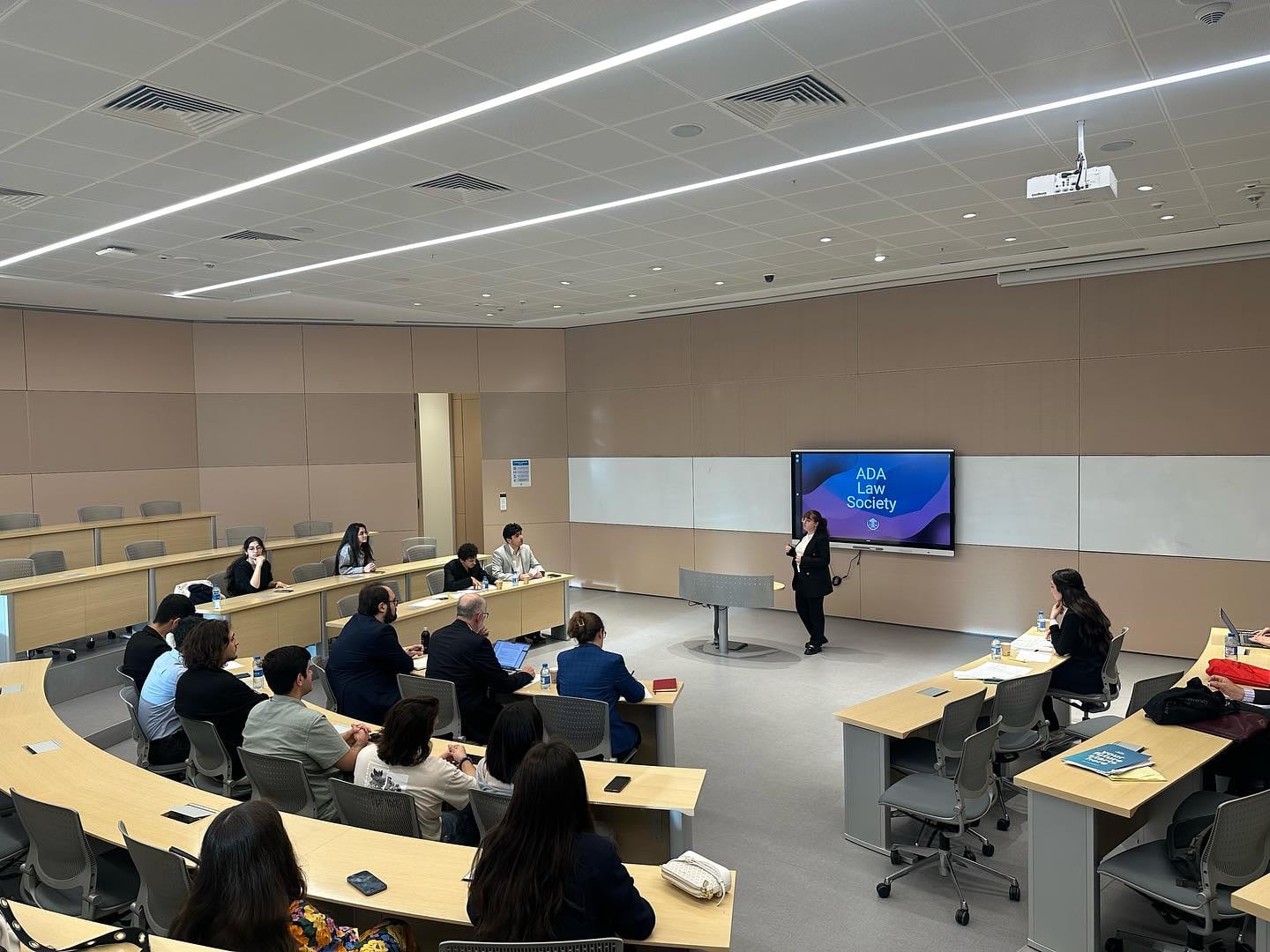A wide lecture hall set out for a debate: a speaker stands in the middle of the floor beneath a screen reading “ADA Law Society”, while students and guests follow from curved desks in the foreground.