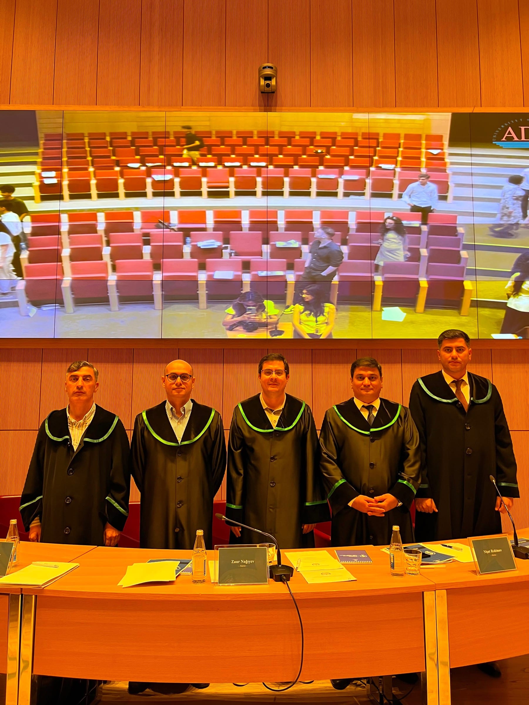 Five judges in academic robes standing in front of the judicial bench, a large screen above them showing the empty auditorium.